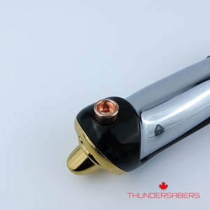 Puede incluir: Primer plano de una empu&ntilde;adura de sable de luz con un dise&ntilde;o negro, dorado y cromado. La empu&ntilde;adura presenta un detalle de color cobre y el texto "THUNDERSABERS" en rojo. El sable de luz tiene una est&eacute;tica elegante y moderna.