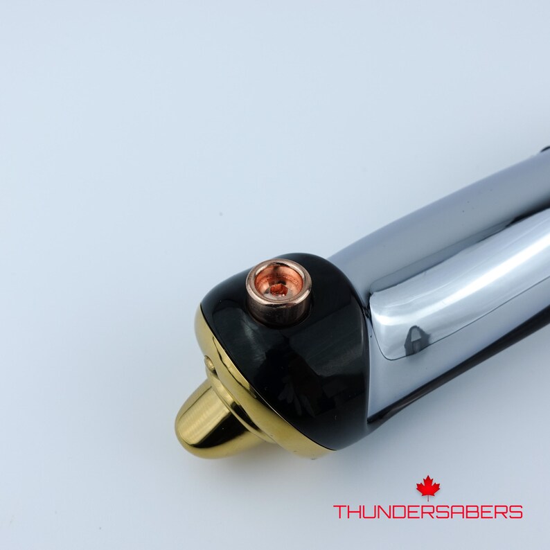 Puede incluir: Primer plano de una empu&ntilde;adura de sable de luz con un dise&ntilde;o negro, dorado y cromado. La empu&ntilde;adura presenta un detalle de color cobre y el texto "THUNDERSABERS" en rojo. El sable de luz tiene una est&eacute;tica elegante y moderna.