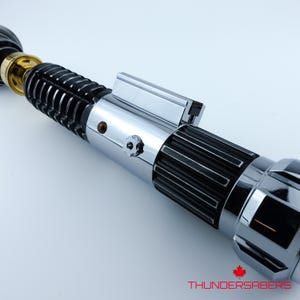 May include: A detailed lightsaber with a silver and black handle, and gold accents. The design includes a series of ridges and a polished finish. The text "THUNDERSABERS" is visible at the bottom.
