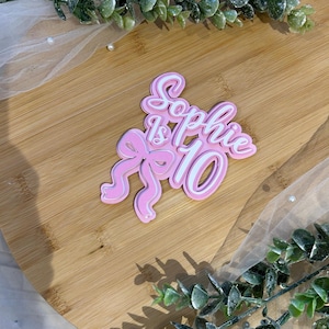 Op de afbeelding: Een roze en witte taarttopper met de tekst "Sophie is 10" in een cursief lettertype. Het getal 10 staat vetgedrukt. De topper heeft een strik ontwerp en ligt op een houten oppervlak.