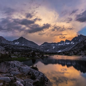 May include: A mountain lake reflecting a colorful sunset sky with clouds. The lake is surrounded by rocky mountains and trees.
