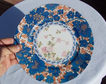 Puede incluir: Un plato de cena de cerámica blanca con un diseño floral azul y dorado. El plato tiene un centro blanco con un patrón floral y un borde azul y dorado.