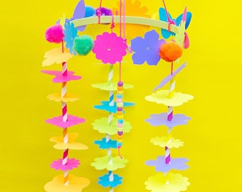 May include: A colorful paper flower mobile with yellow, pink, blue, and purple flowers hanging from a wooden ring. The flowers are strung on colorful straws and beads.