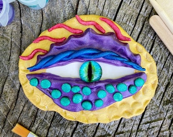 May include: A close-up of a colorful clay eye with a green iris and purple eyelids. The eye is surrounded by a yellow background with red and pink accents.