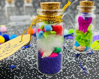 May include: A small glass bottle filled with colorful beads, glitter, and a seashell. The bottle has a cork stopper and is decorated with a gold ribbon.