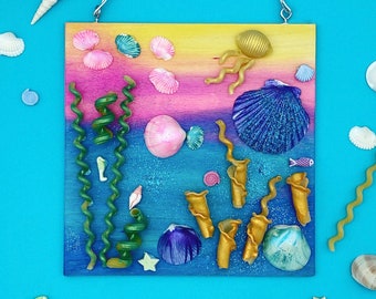 May include: Square wooden wall art featuring an underwater scene with seashells, seaweed, and gold-colored coral. The background transitions from yellow to pink to blue. A wire is attached for hanging. Additional seashells are scattered around the artwork.