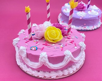 May include: Two decorative cakes on a pink background. The front cake is pink with white frosting, decorative flowers, sprinkles, and striped candles. The second cake is purple with white frosting, a rose, and candles. A festive, celebratory theme.