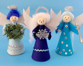 May include: Three felt angel ornaments with wings, each with a different color dress and hair. One angel holds a green wreath, one holds a silver snowflake, and one holds a silver trumpet.