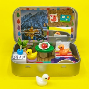 May include: A miniature diorama inside a silver tin, featuring a landscape scene, a small plant, and several toy ducks. The scene includes a miniature bed, table, and other decorative elements, all set against a yellow background.