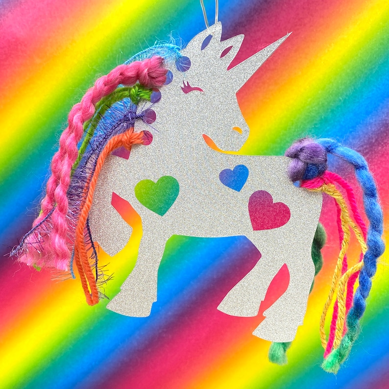 May include: A silver glitter unicorn cutout with a rainbow yarn mane and tail. The unicorn has three hearts on its body, one green, one pink, and one blue.