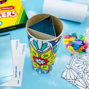 May include: A handmade kaleidoscope with a colorful floral design. The kaleidoscope is made from a cardboard tube and features a triangular prism inside. Additional craft supplies, including markers and labels, are visible.