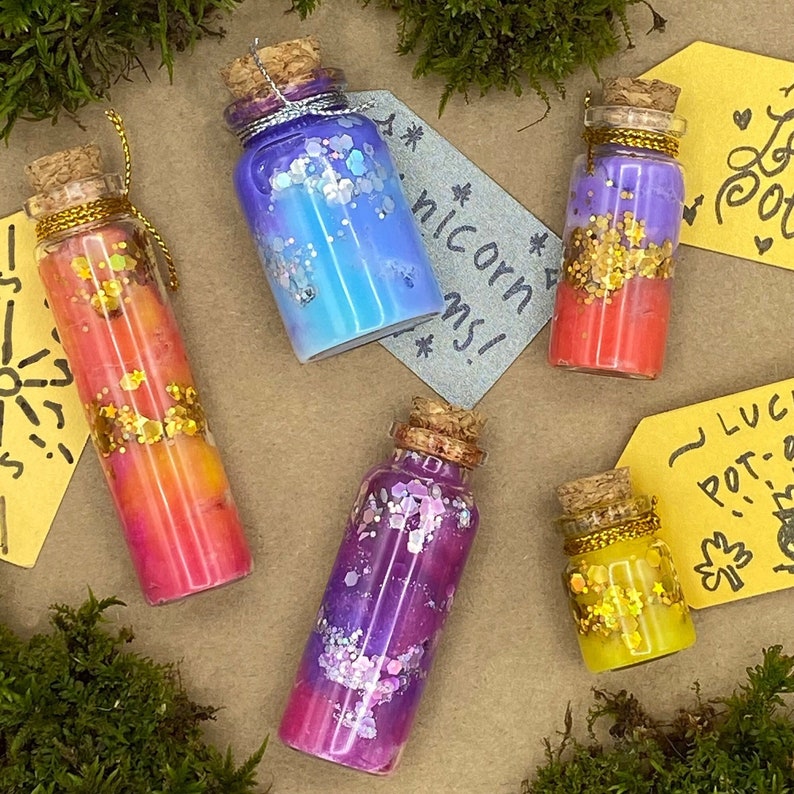 May include: Five small glass bottles with cork stoppers, filled with colorful liquids and glitter. The bottles are decorated with gold and silver accents. The background includes moss and handwritten tags with text.