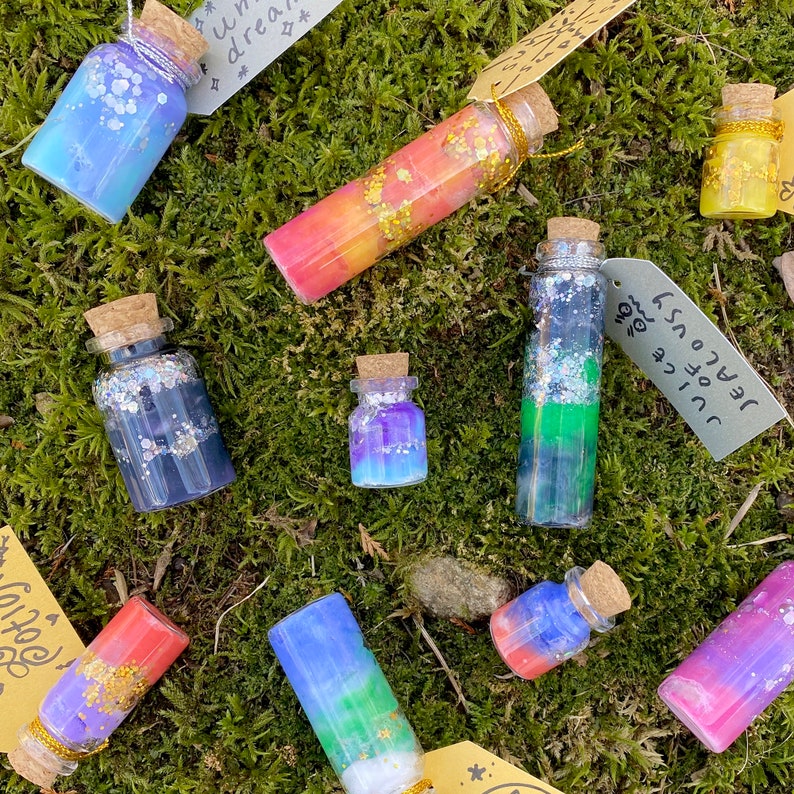 May include: A collection of small glass bottles with cork stoppers, filled with colorful liquids and glitter. The bottles are arranged on a bed of green moss, with small tags attached. Some bottles have blue, purple, and yellow hues.