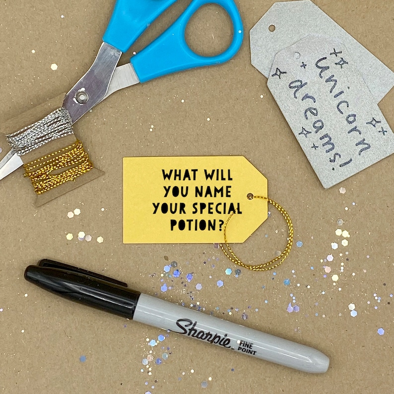 May include: A craft scene with a yellow tag that reads "WHAT WILL YOU NAME YOUR SPECIAL POTION?" and a silver tag that reads "unicorn dreams!". Includes blue scissors, a black Sharpie, and gold and silver string.