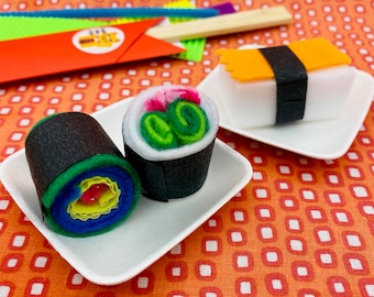 May include: Two felt sushi rolls on white plates. One roll is green, yellow, blue, and red. The other roll is green, white, and black. Both rolls have black seaweed on the outside.