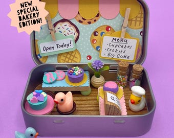 May include: A miniature bakery scene inside a metal tin. The scene includes tiny cakes, cookies, and a duck figurine. The sign says "Open Today!" and "Menu: Cupcakes, Cookies, Big Cake." A starburst sign reads "New Special Bakery Edition!"