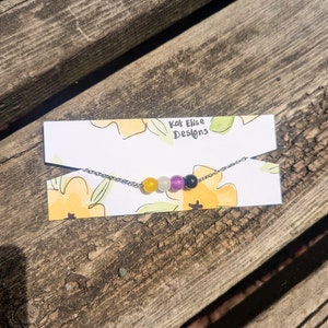 May include: A silver chain necklace with five gemstone beads in the colors of the asexual pride flag: black, white, purple, gray, and yellow. The necklace is on a white card with the text "Kat Elise Designs".