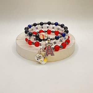 May include: Stack of three beaded bracelets with red, black, blue, and clear beads. A charm with the letter "A" and a baseball hangs from the bracelets. The bracelets are displayed on a round, light-colored platform.