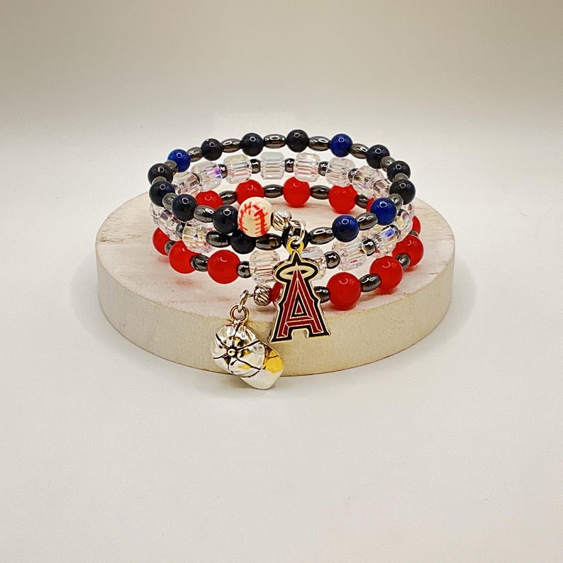 May include: Stack of three beaded bracelets with red, black, blue, and clear beads. A charm with the letter "A" and a baseball hangs from the bracelets. The bracelets are displayed on a round, light-colored platform.