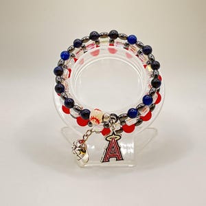 May include: Stacked beaded bracelets with red, blue, and black beads. A baseball charm and a Los Angeles Angels logo charm dangle from the bracelets. The bracelets are displayed on a clear, circular stand.