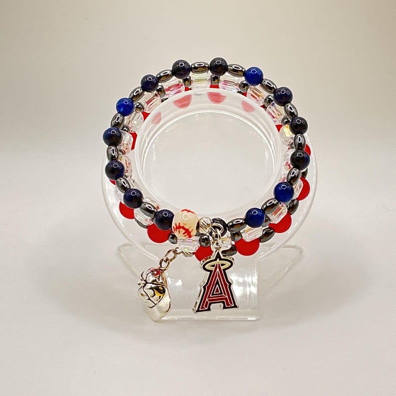 May include: Stacked beaded bracelets with red, blue, and black beads. A baseball charm and a Los Angeles Angels logo charm dangle from the bracelets. The bracelets are displayed on a clear, circular stand.