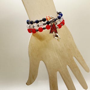 May include: A stack of three beaded bracelets in red, white, and blue. The bracelets have a baseball charm and a red and white "A" charm. The bracelets are on a wooden hand-shaped display.