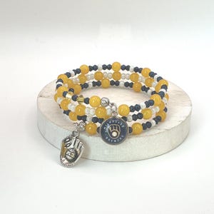 May include: A multi-strand wrap bracelet featuring alternating yellow, white, and dark blue beads. The bracelet includes two silver-toned metal charms: one shaped like a baseball glove and a circular charm displaying the Milwaukee Brewers logo.