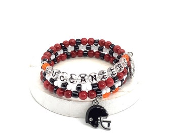 Puede incluir: Un brazalete de cuentas de múltiples hilos con cuentas rojas, blancas y negras. El brazalete tiene un dije de casco de fútbol y la palabra "BUCCANEERS" deletreada en cuentas transparentes.