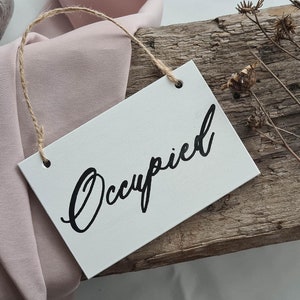 May include: A white sign with the word "Occupied" written in black cursive script. The sign is hanging from a piece of twine and is resting on a piece of weathered wood.