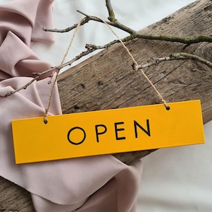 Peut inclure: Une enseigne en bois jaune avec le mot "OUVERT" en lettres noires est suspendue à un morceau de ficelle. L'enseigne repose sur un morceau de bois usé avec une branche de brindilles et un morceau de tissu rose.