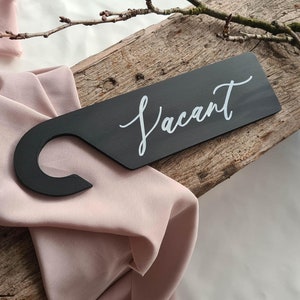 May include: Black wooden door hanger with a white cursive inscription that reads "Vacant". The hanger has a hook at the top.