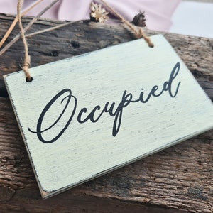 May include: A rustic wooden sign with a distressed, light green paint finish. The sign is hanging from a string and reads "Occupied" in black cursive lettering.