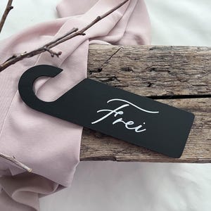 May include: A black door hanger with the word "Frei" in white script. The hanger is rectangular with a curved top for hanging. It rests on a wooden surface with a pink fabric and twigs.