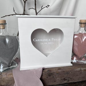 May include: A white wooden box with a heart-shaped cutout, personalized with the names "Caroline & Philip" and the date "14.06.2025". Flanking the box are two heart-shaped glass bottles filled with gray and pink sand, respectively. A decorative branch sits atop the box.