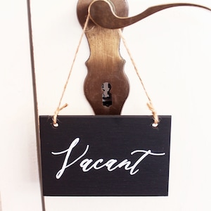 May include: A black chalkboard sign with white lettering that says "Vacant" hanging on a door knob.