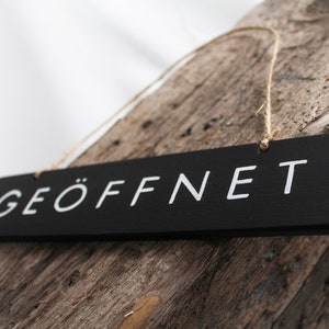以下が含まれることがあります： ひもで吊るされた、白文字で「GEÖFFNET」と書かれた黒い木製の看板です。