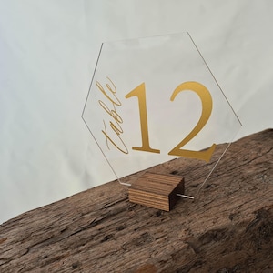 May include: Clear acrylic table number 12 with gold lettering. The hexagonal sign is mounted on a small wooden base. The word "Table" is written in cursive above the number 12. The sign is displayed on a rustic wooden surface.