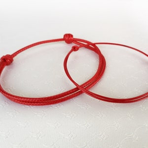 Puede incluir: Dos pulseras de cuerda roja atadas en un nudo. Las pulseras están hechas de cuerda roja fina.