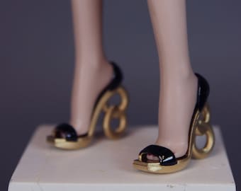 Puede incluir: Sandalias de tacón alto negras y doradas. Las sandalias presentan una base dorada, correas negras y un tacón dorado distintivo con forma de letra estilizada. El diseño de punta abierta añade un toque de elegancia.