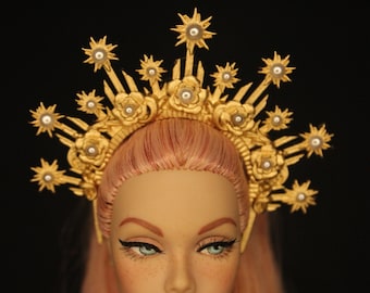 May include: A doll with pink hair wearing a gold crown with pearl accents. The crown is made of metal and has a starburst design.