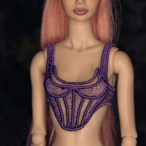 May include: A doll with pink hair styled in a ponytail with a gold hair clip. The doll is wearing a purple lace corset top.