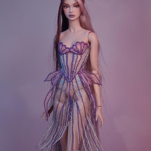 May include: A fashion doll wearing a strapless, intricately designed gown. The dress features a purple and white color scheme with a seashell-shaped bodice and flowing skirt. The doll has long, flowing hair and is posed against a soft, purple background.