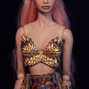 May include: A doll with long pink hair styled in a ponytail, wearing a gold feathered bra and a colorful patterned skirt.