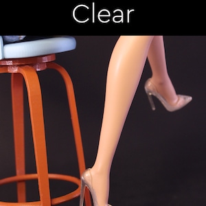 Puede incluir: Un par de zapatos de tac&oacute;n alto transparentes con punta puntiaguda. Los zapatos son usados por una persona sentada en un taburete. La palabra "Claro" est&aacute; escrita en blanco sobre un fondo negro.