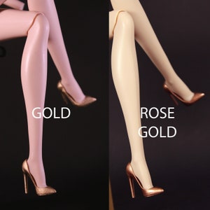 Puede incluir: Dos pares de zapatos de tacón alto con punta. Los zapatos son del mismo estilo pero un par es dorado y el otro es dorado rosa. Los zapatos están en las piernas de una muñeca.