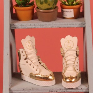 May include: A pair of white leather high-top sneakers with gold accents and an anchor logo on the side. The sneakers are on a gray shelf with a pink background.