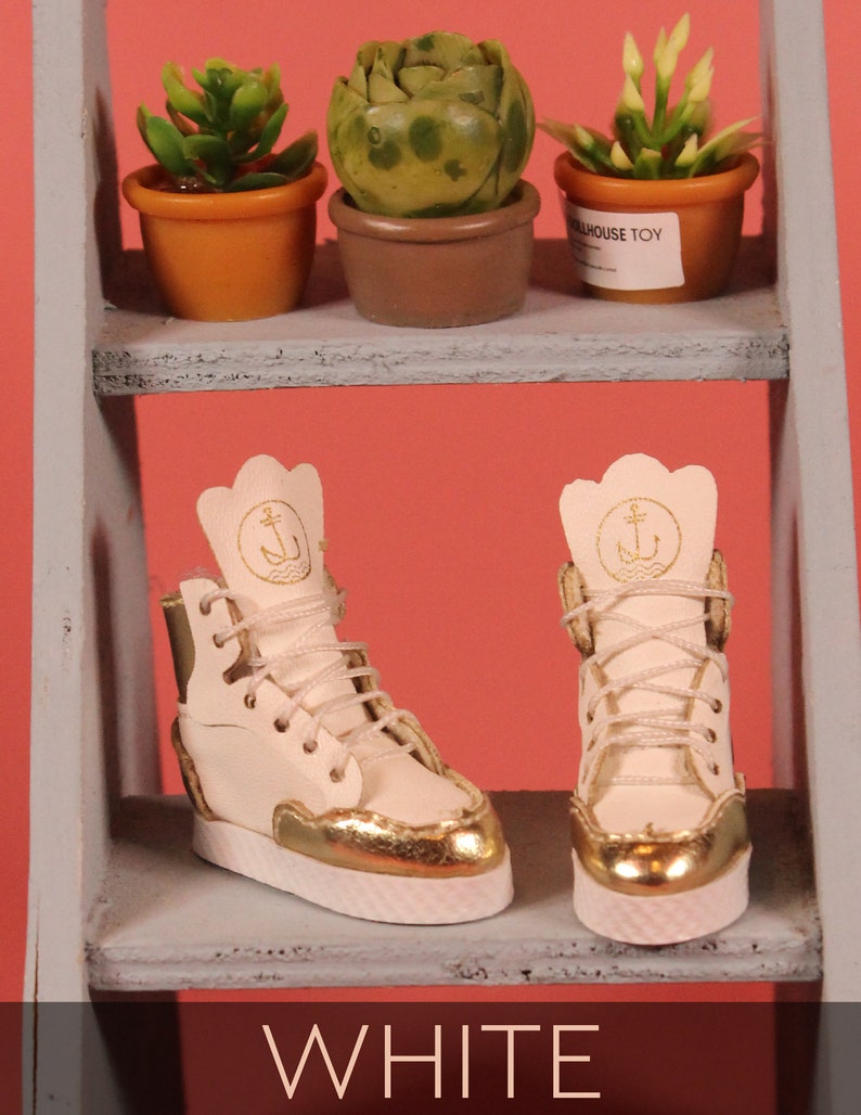 May include: A pair of white leather high-top sneakers with gold accents and an anchor logo on the side. The sneakers are on a gray shelf with a pink background.