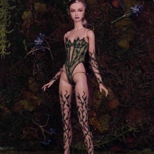 May include: A fashion doll wearing a green bodysuit and leggings with a leaf pattern. The doll has brown hair and is set against a mossy backdrop. The bodysuit features a leaf-shaped neckline.