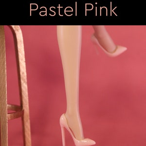 Puede incluir: Un par de zapatos de tac&oacute;n alto de color rosa pastel. Los zapatos est&aacute;n hechos de un material liso y brillante y tienen una punta puntiaguda. Los zapatos son usados por las piernas de una mu&ntilde;eca, que tambi&eacute;n son de color rosa pastel.
