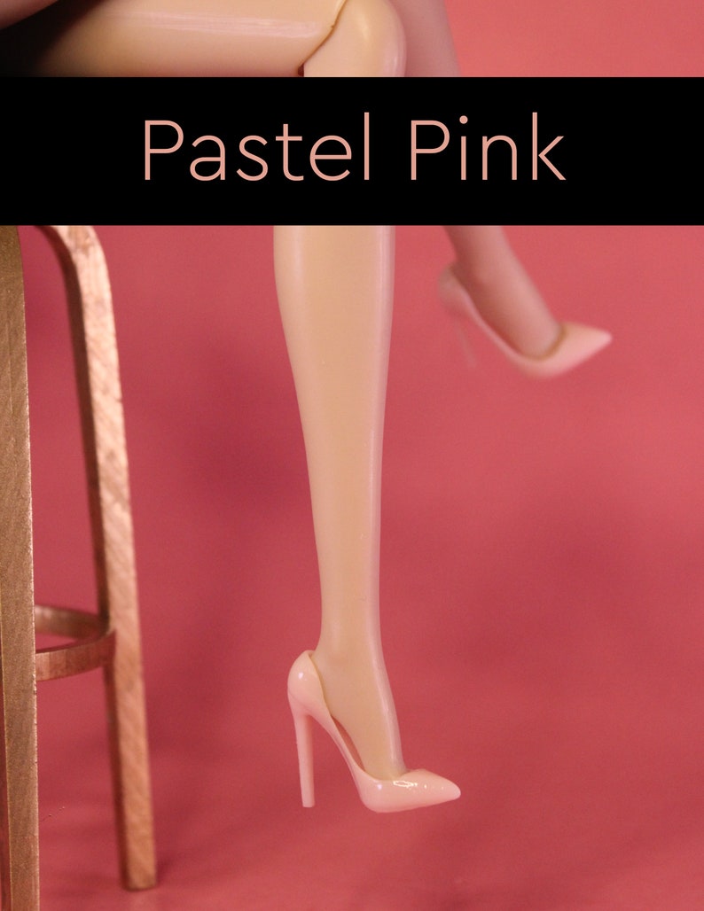 Puede incluir: Un par de zapatos de tac&oacute;n alto de color rosa pastel. Los zapatos est&aacute;n hechos de un material liso y brillante y tienen una punta puntiaguda. Los zapatos son usados por las piernas de una mu&ntilde;eca, que tambi&eacute;n son de color rosa pastel.